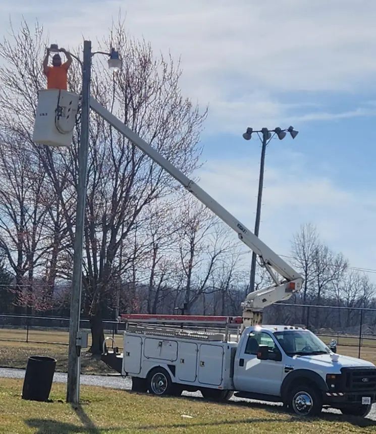 Lineman in a bucket truck working on a pole light beside a fenced ball field